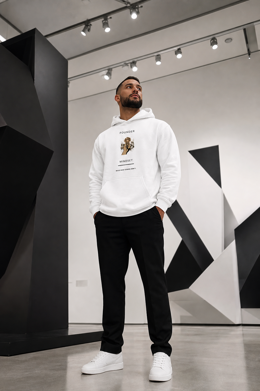 A man wearing a white "Founder Mindset" graphic hoodie, black pants, and white sneakers standing in a modern studio with geometric black-and-white background architecture.